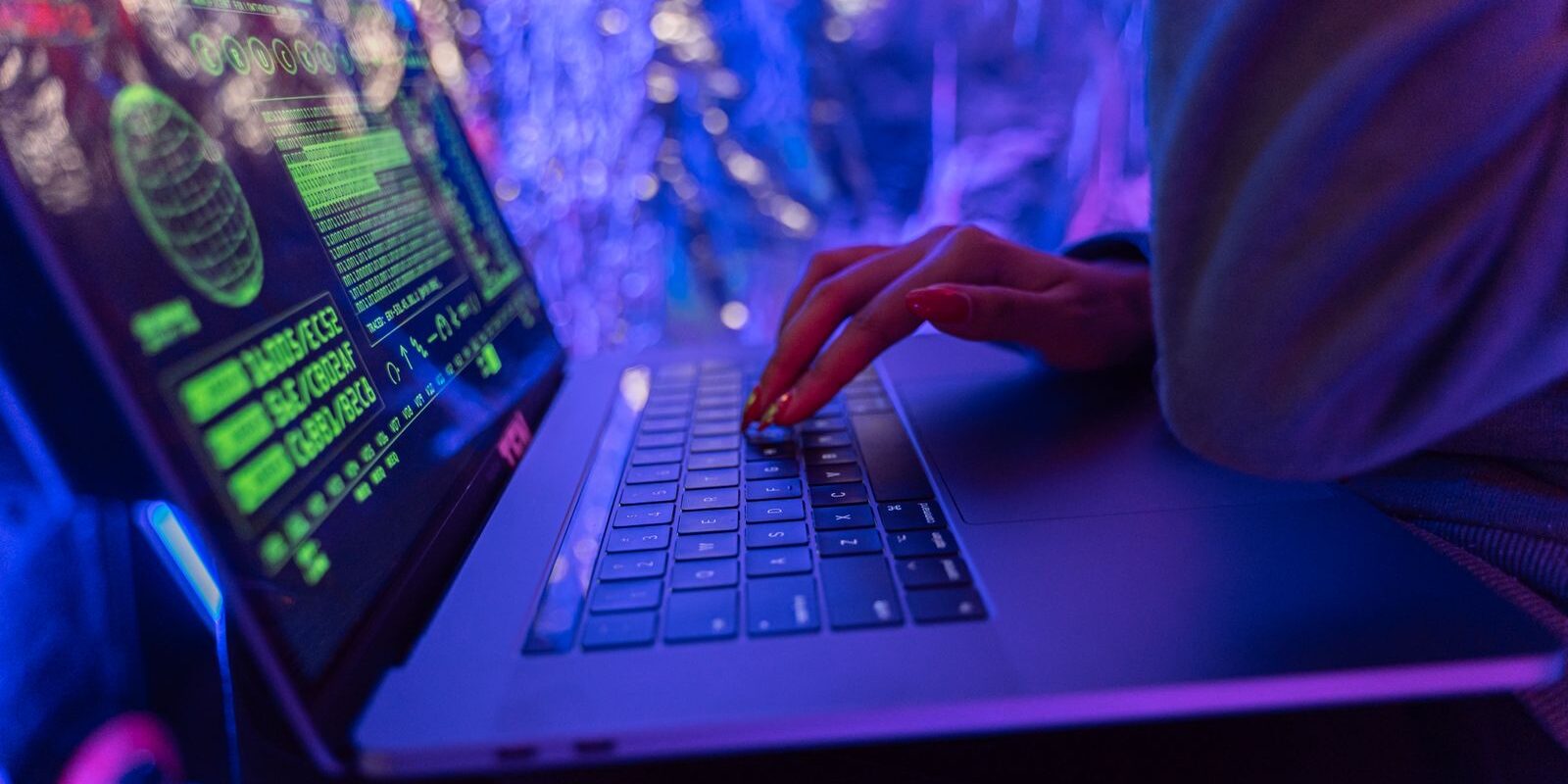Manos de una persona escribiendo en una laptop con gráficos de ciberseguridad en la pantalla, bajo luces de neón azules y moradas