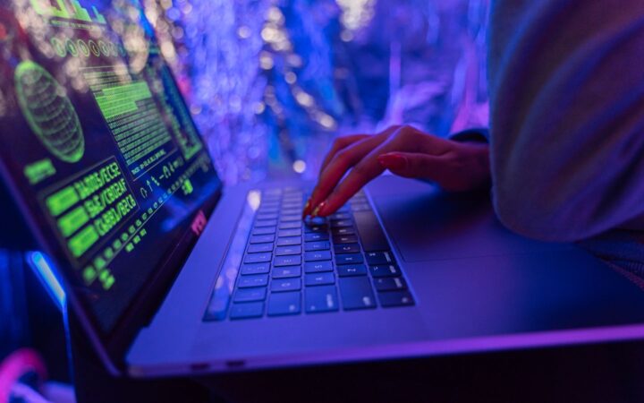 Manos de una persona escribiendo en una laptop con gráficos de ciberseguridad en la pantalla, bajo luces de neón azules y moradas