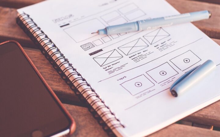 Cuaderno con bocetos a mano del diseño de una página web, junto a una pluma y un celular sobre una mesa de madera