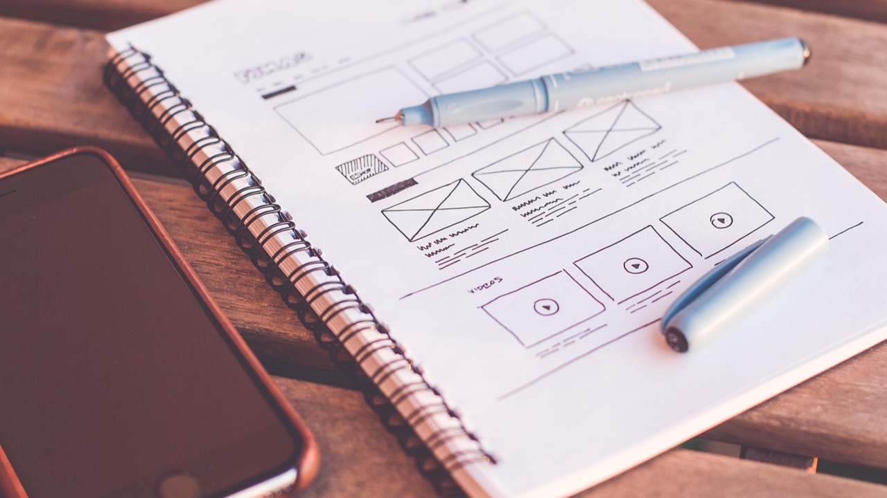 Cuaderno con bocetos a mano del diseño de una página web, junto a una pluma y un celular sobre una mesa de madera