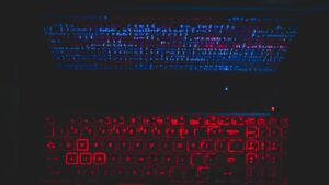 Laptop en un cuarto oscuro con el teclado iluminado en rojo y líneas de código reflejadas en azul
