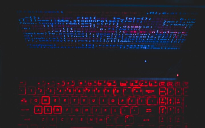 Laptop en un cuarto oscuro con el teclado iluminado en rojo y líneas de código reflejadas en azul
