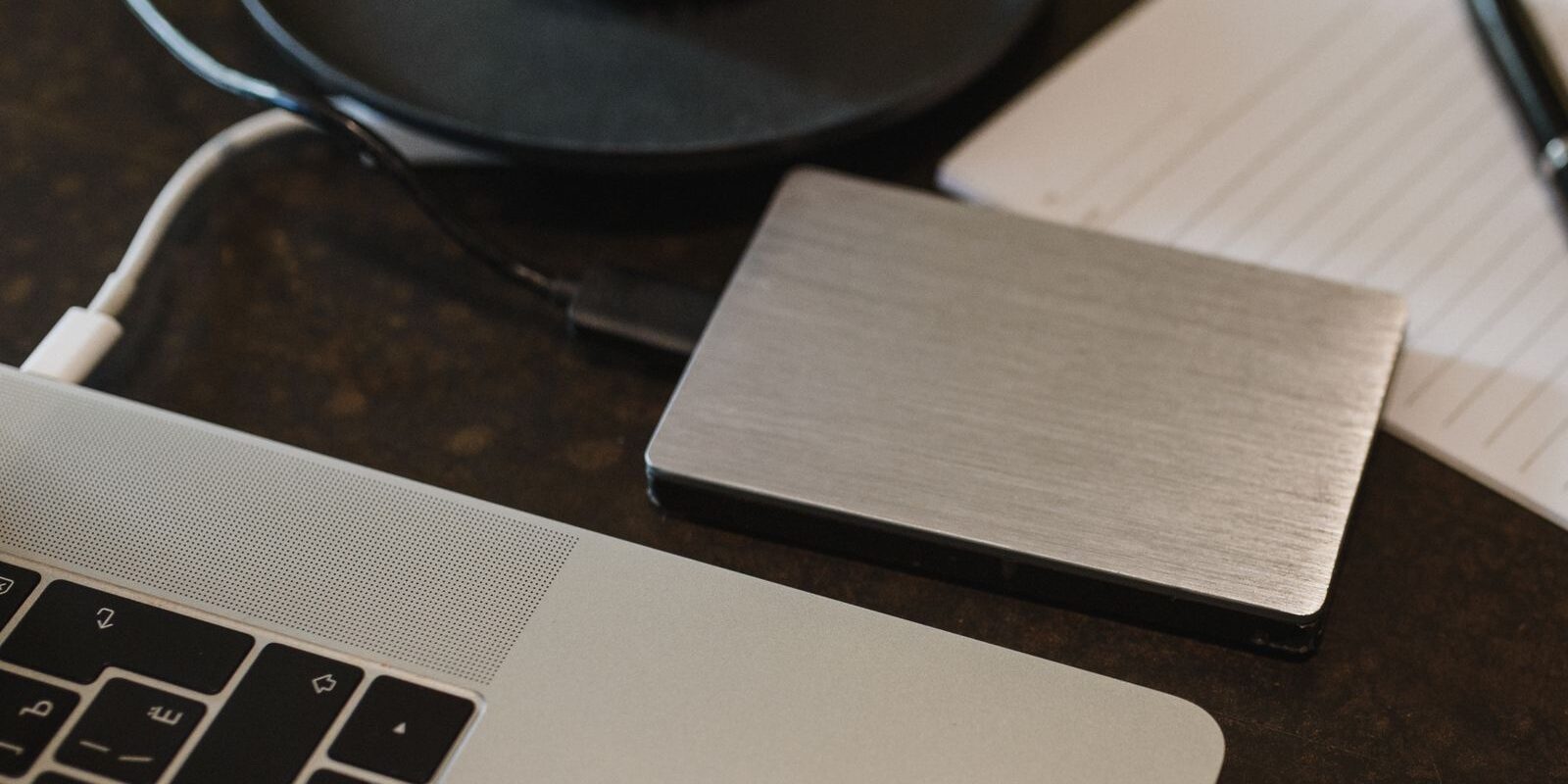 Disco duro externo plateado conectado con un cable a una laptop sobre un escritorio oscuro