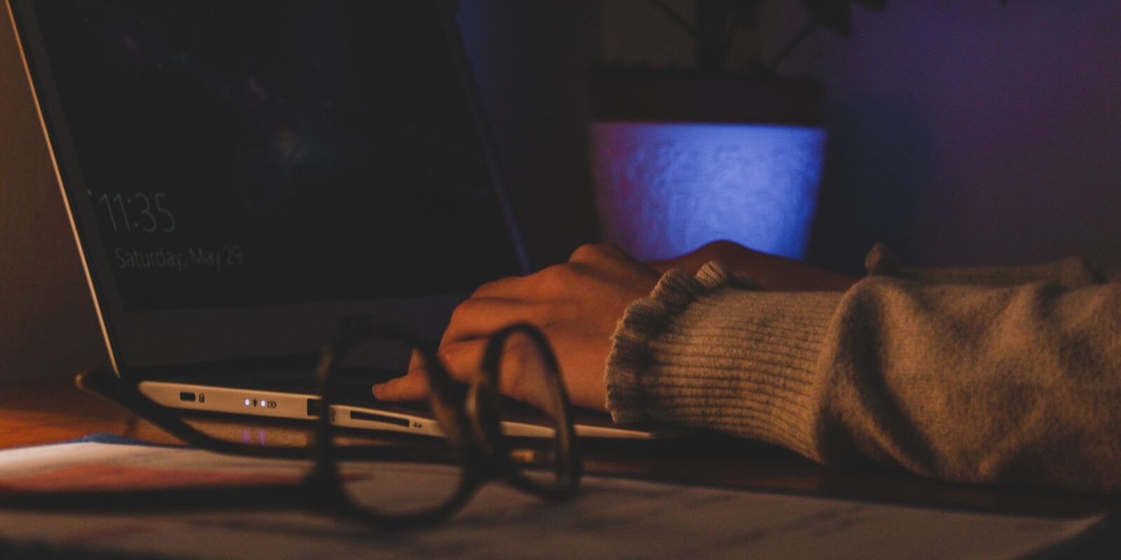 Manos escribiendo en una laptop en un cuarto con poca luz, con unos lentes sobre el escritorio