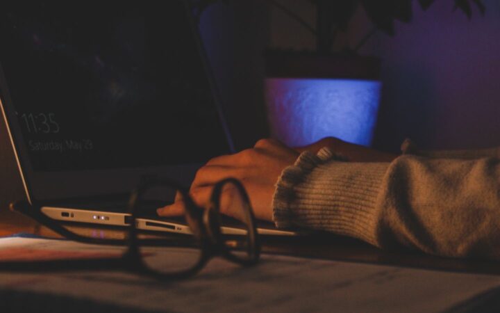 Manos escribiendo en una laptop en un cuarto con poca luz, con unos lentes sobre el escritorio