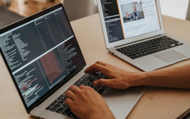 Manos de una persona escribiendo en una laptop con una terminal abierta junto a otra laptop sobre una mesa de madera