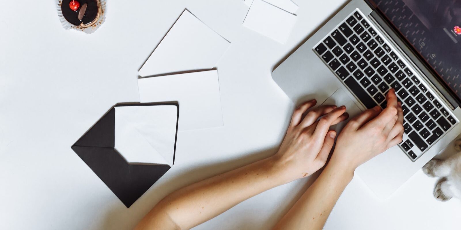 Manos de una persona escribiendo en una laptop sobre un escritorio blanco con sobres y hojas de papel
