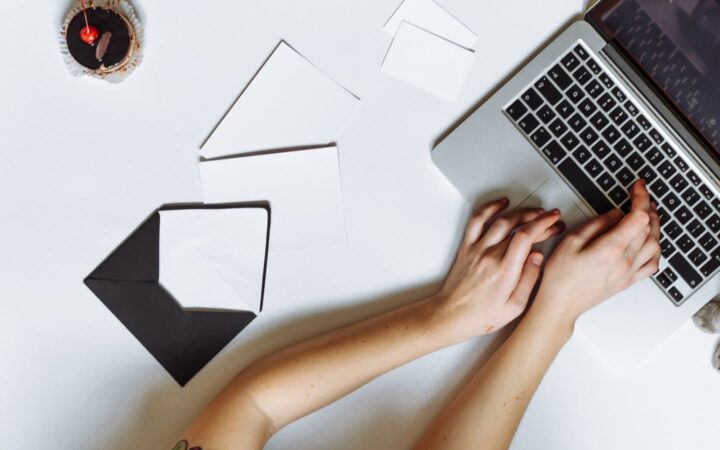 Manos de una persona escribiendo en una laptop sobre un escritorio blanco con sobres y hojas de papel