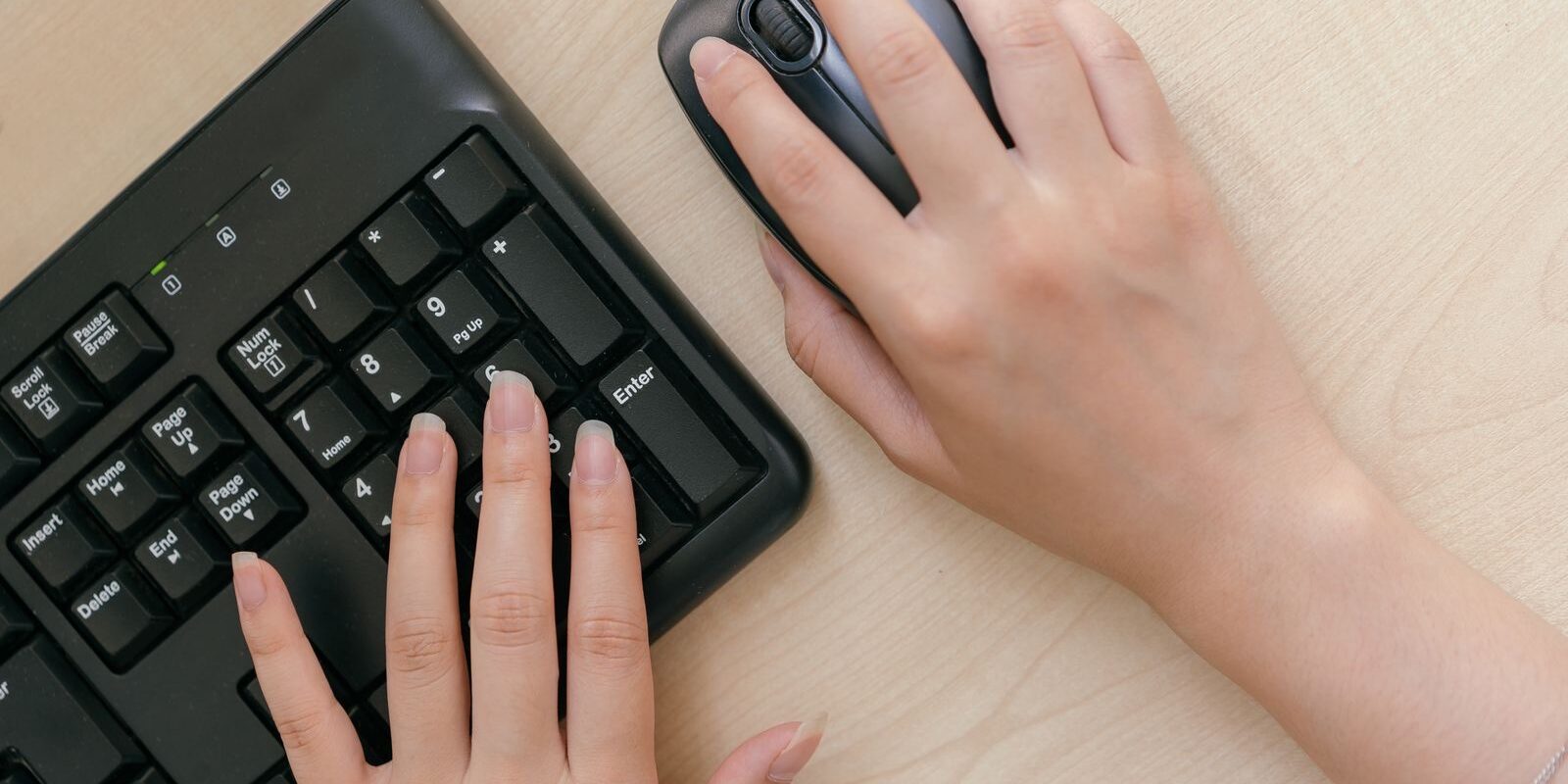 Manos de una persona usando un teclado negro y un mouse sobre un escritorio de madera clara