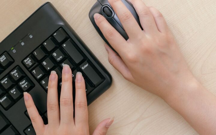 Manos de una persona usando un teclado negro y un mouse sobre un escritorio de madera clara