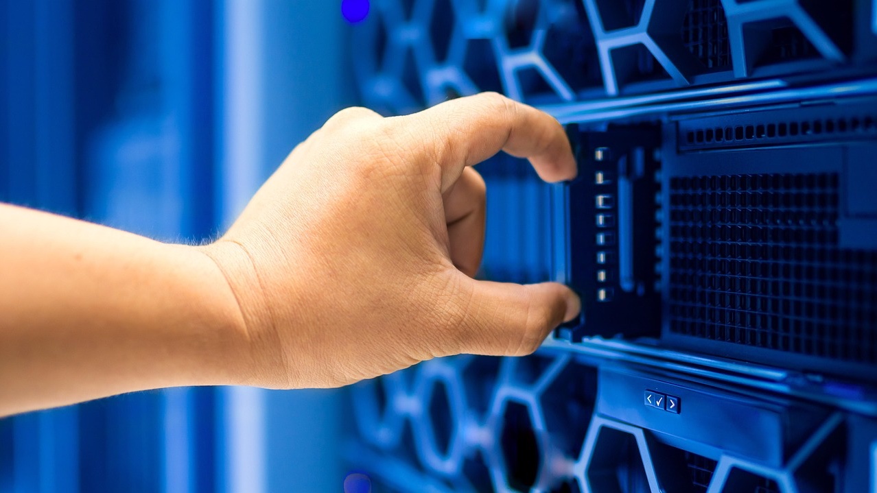 Mano de una persona tomando un servidor de un rack iluminado en azul dentro de un centro de datos