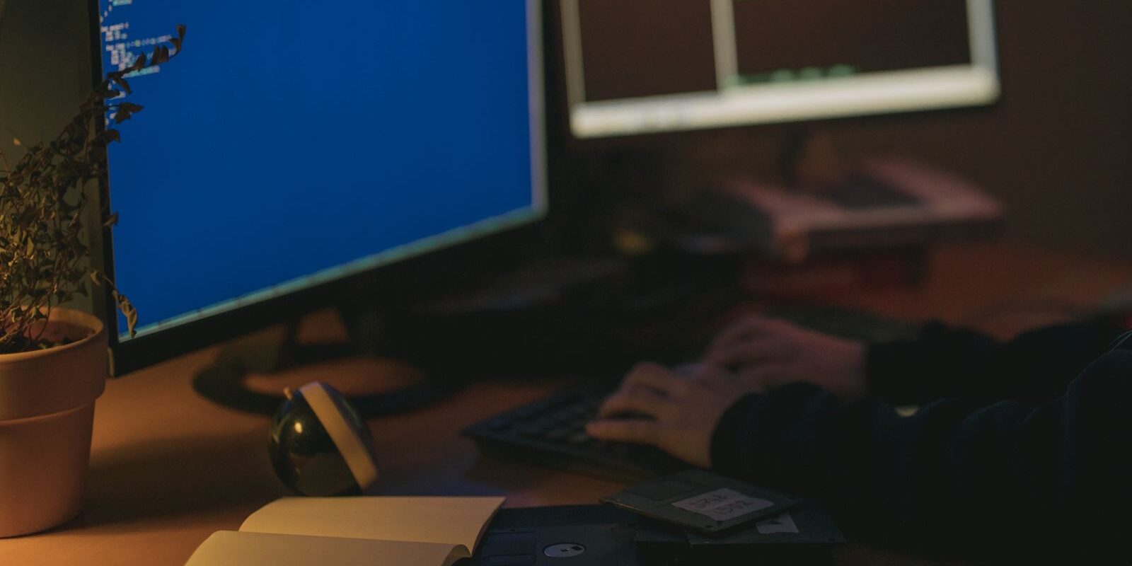 Persona escribiendo en un teclado frente a dos monitores en una habitación con poca luz