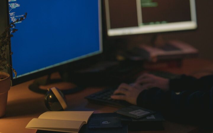 Persona escribiendo en un teclado frente a dos monitores en una habitación con poca luz