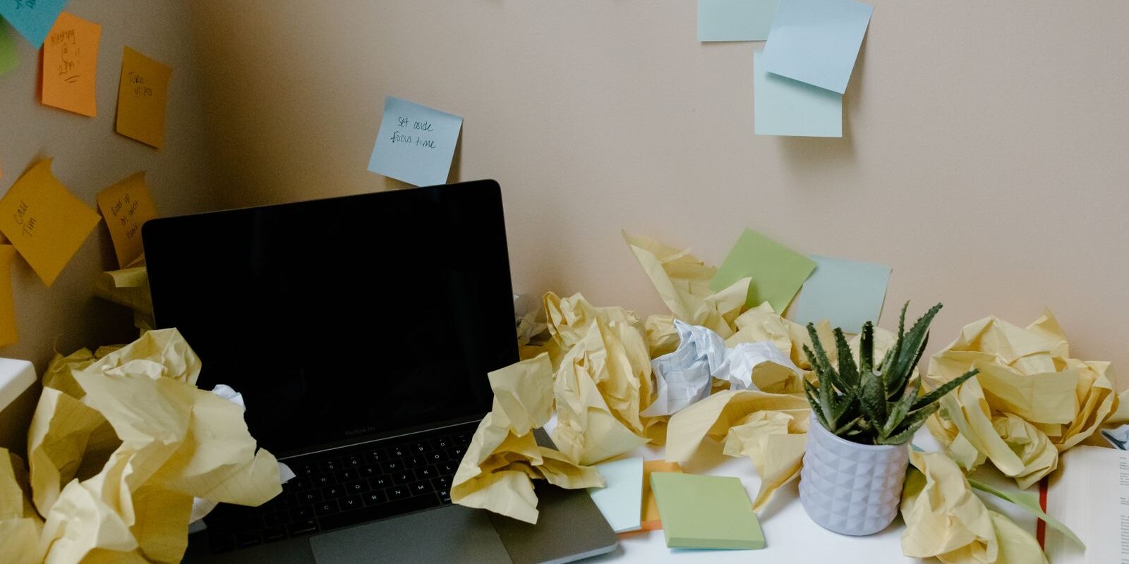Laptop sobre un escritorio rodeada de bolas de papel arrugado y notas adhesivas pegadas en la pared