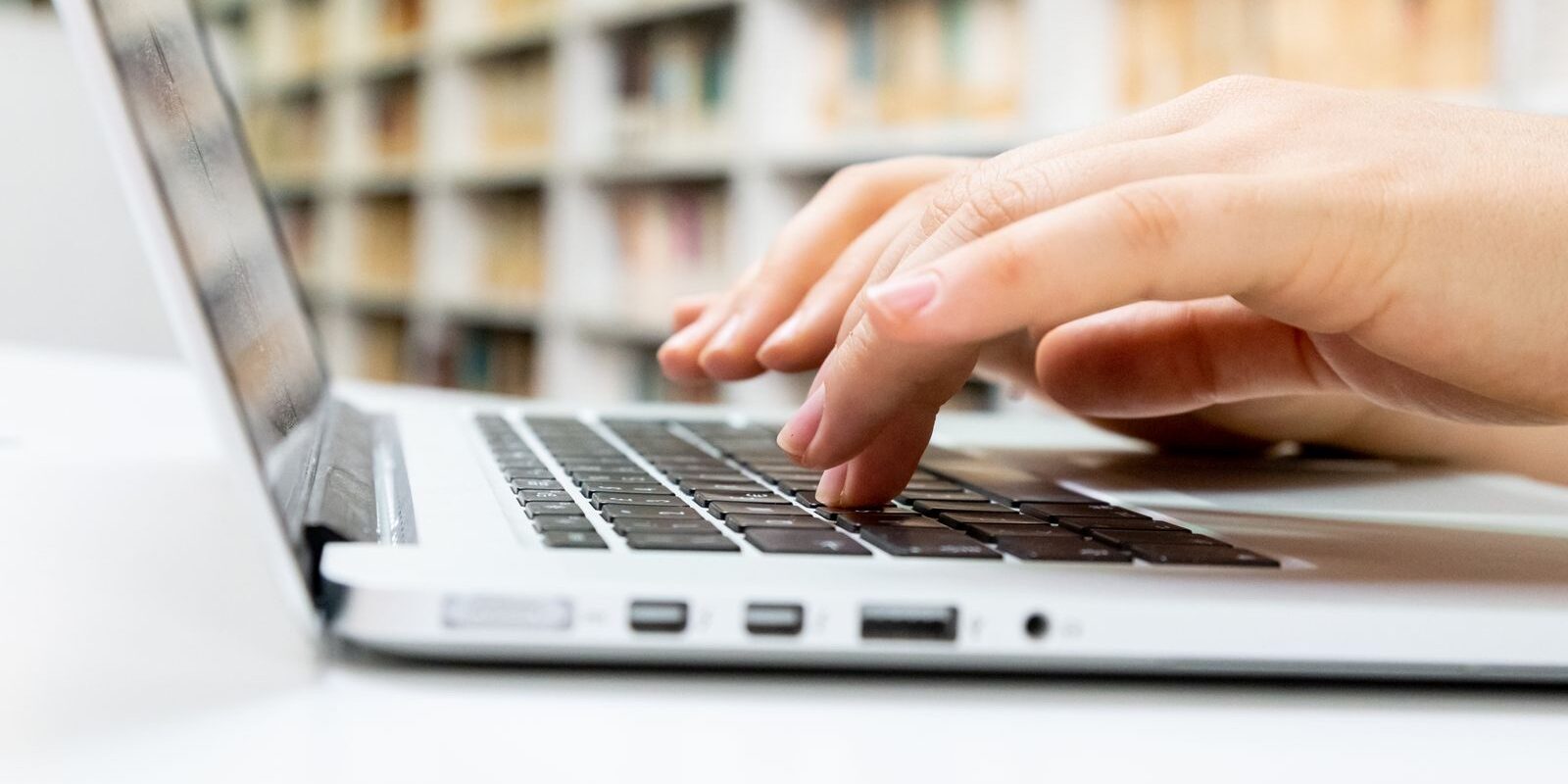 Manos de una persona escribiendo en el teclado de una laptop plateada dentro de una biblioteca
