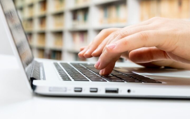 Manos de una persona escribiendo en el teclado de una laptop plateada dentro de una biblioteca