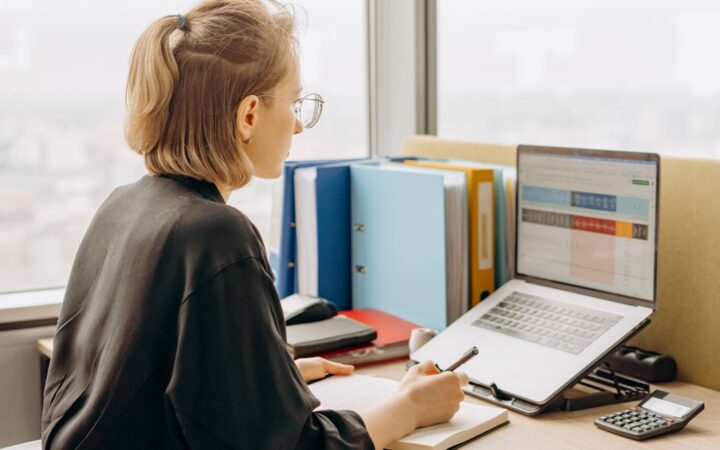 Mujer con lentes revisa una hoja de cálculo en una laptop mientras toma notas en una oficina