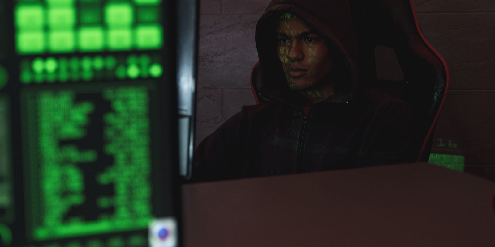Persona con sudadera con capucha frente a una computadora en un cuarto oscuro iluminado por una pantalla verde