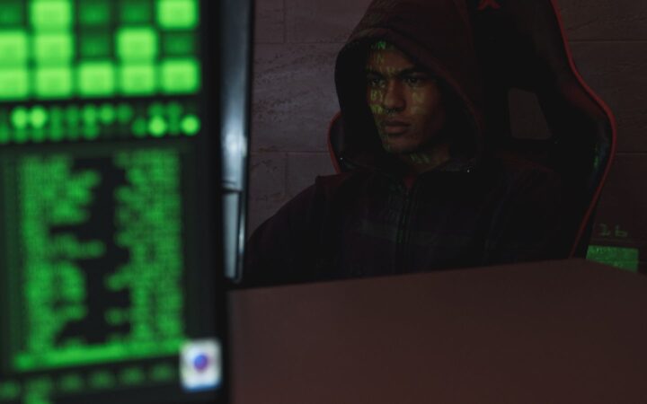 Persona con sudadera con capucha frente a una computadora en un cuarto oscuro iluminado por una pantalla verde