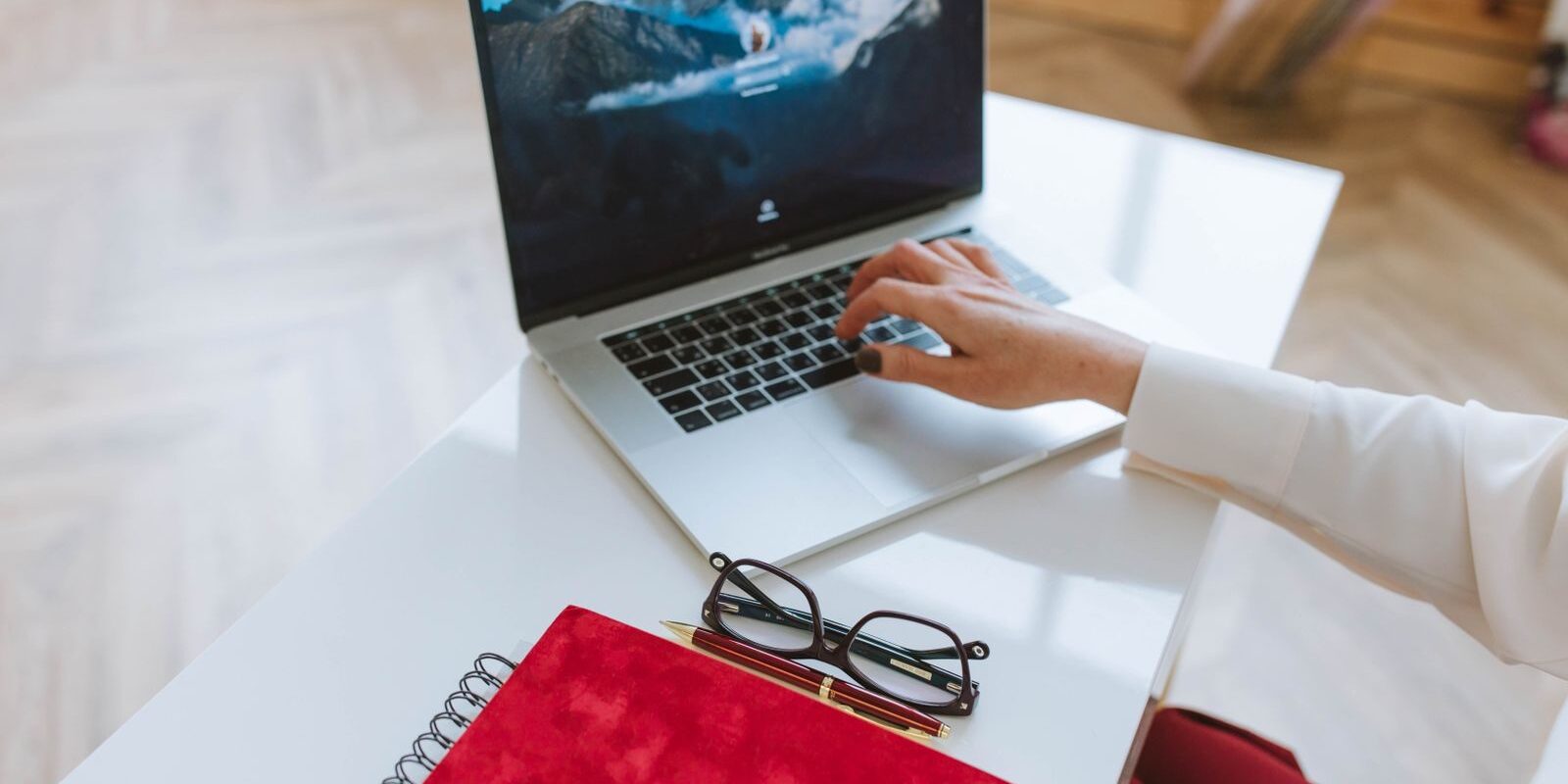 Mano de una persona escribiendo en una laptop sobre un escritorio blanco junto a una libreta roja y unos lentes