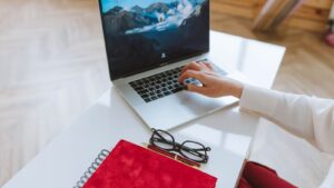 Mano de una persona escribiendo en una laptop sobre un escritorio blanco junto a una libreta roja y unos lentes