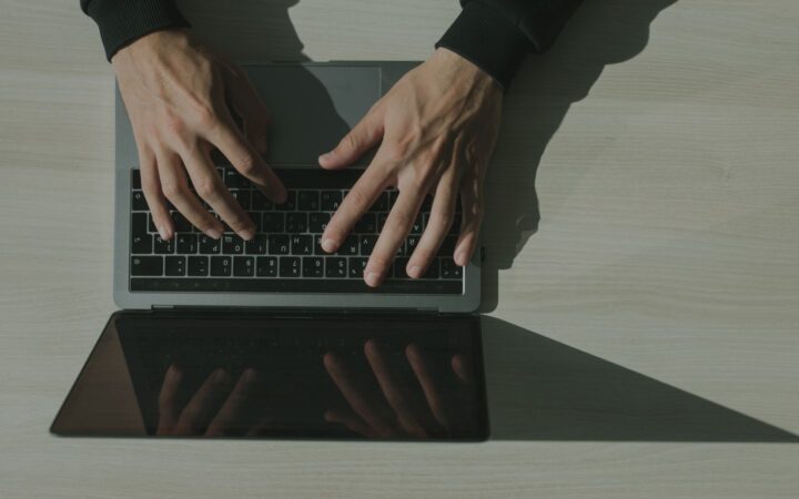 Manos de una persona escribiendo en una laptop sobre un escritorio claro, vista desde arriba