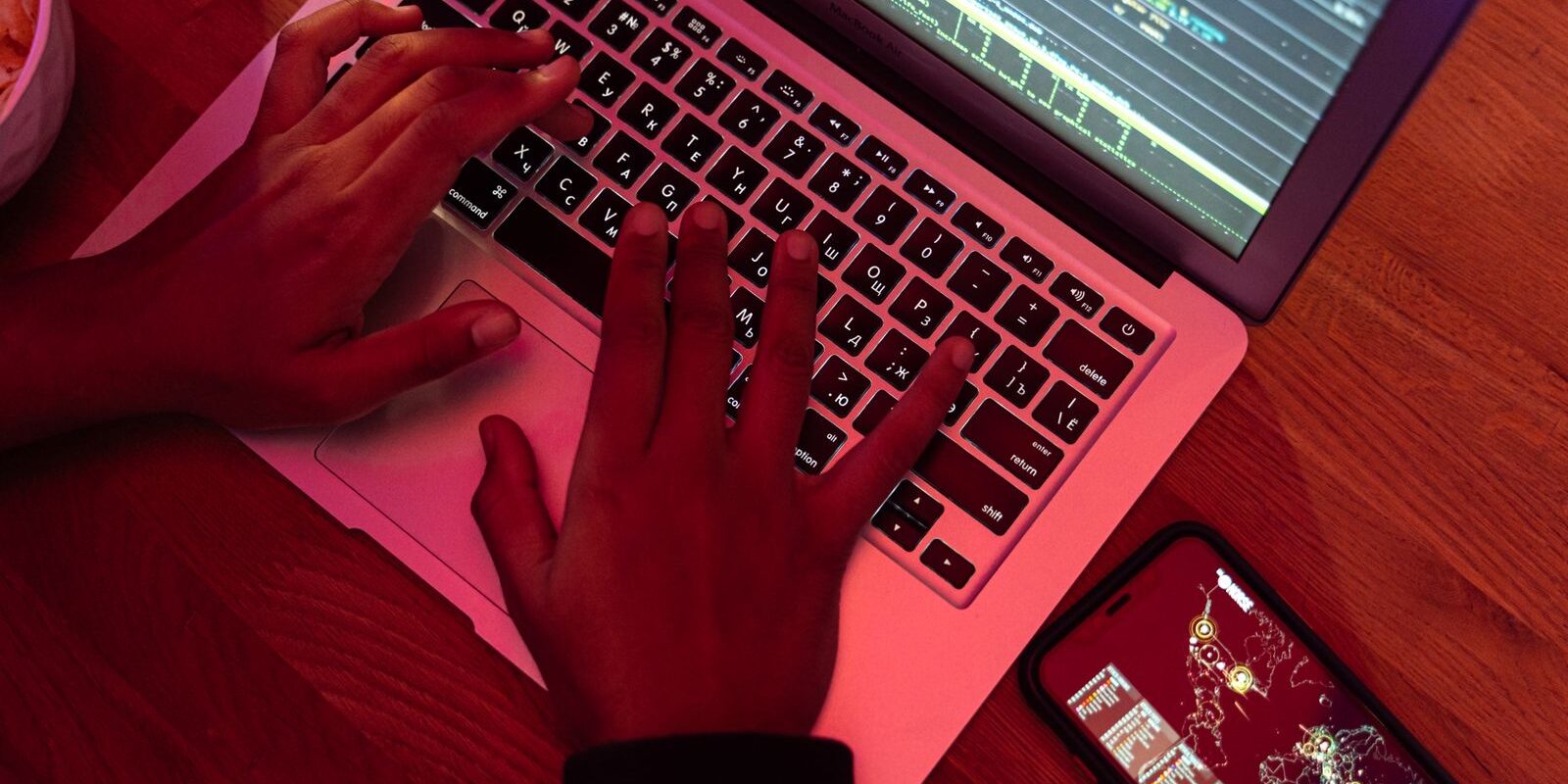 Manos escribiendo en una laptop con código en pantalla bajo una luz rojiza, junto a un celular