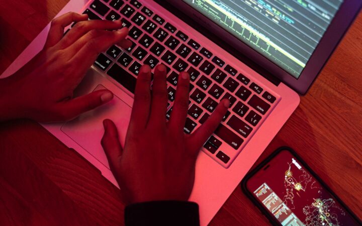 Manos escribiendo en una laptop con código en pantalla bajo una luz rojiza, junto a un celular