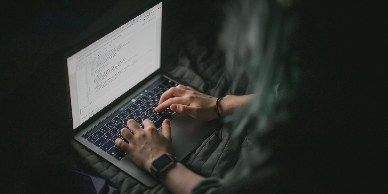 Manos de una persona escribiendo en una laptop con código en pantalla dentro de una habitación oscura