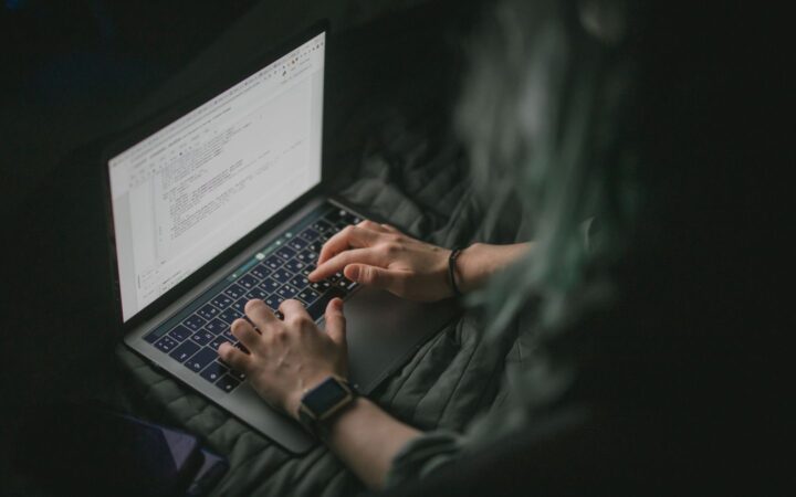 Manos de una persona escribiendo en una laptop con código en pantalla dentro de una habitación oscura