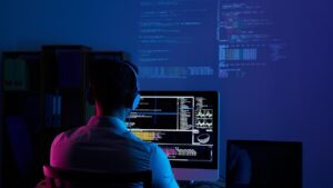 Programador de espaldas con audífonos trabajando frente a un monitor en una oficina con luz azul