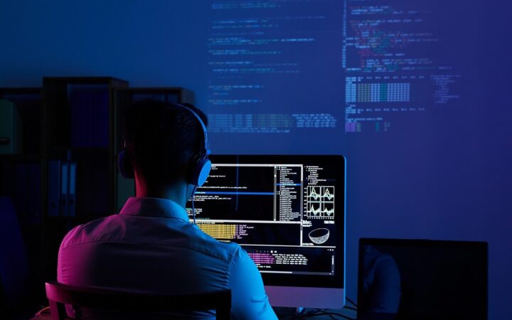 Programador de espaldas con audífonos trabajando frente a un monitor en una oficina con luz azul