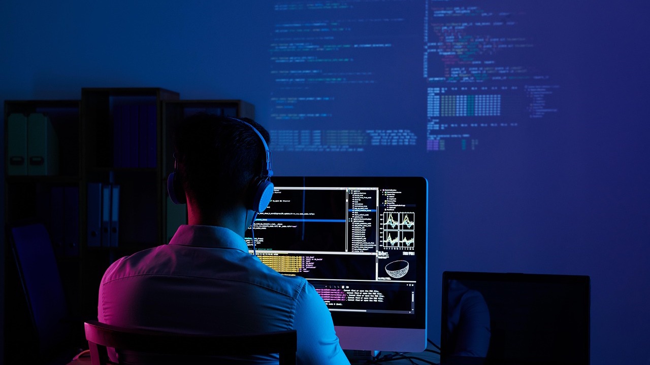 Programador de espaldas con audífonos trabajando frente a un monitor en una oficina con luz azul