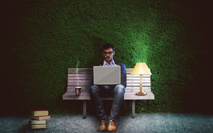 Un hombre trabaja con su laptop sentado en una banca junto a una lámpara y una taza de café